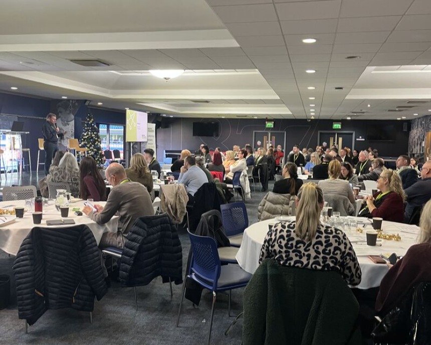 Conference room with round tables, people seated, and a speaker presenting near a Christmas tree