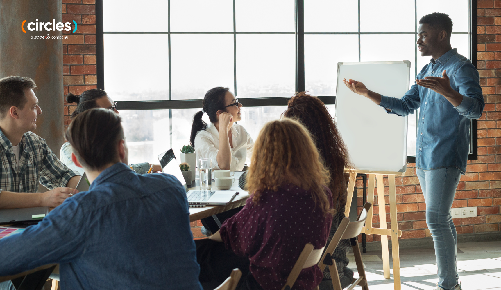 A professional team meeting in a sunlit industrial-style office. A man stands at a whiteboard presenting to a group of seated coworkers.