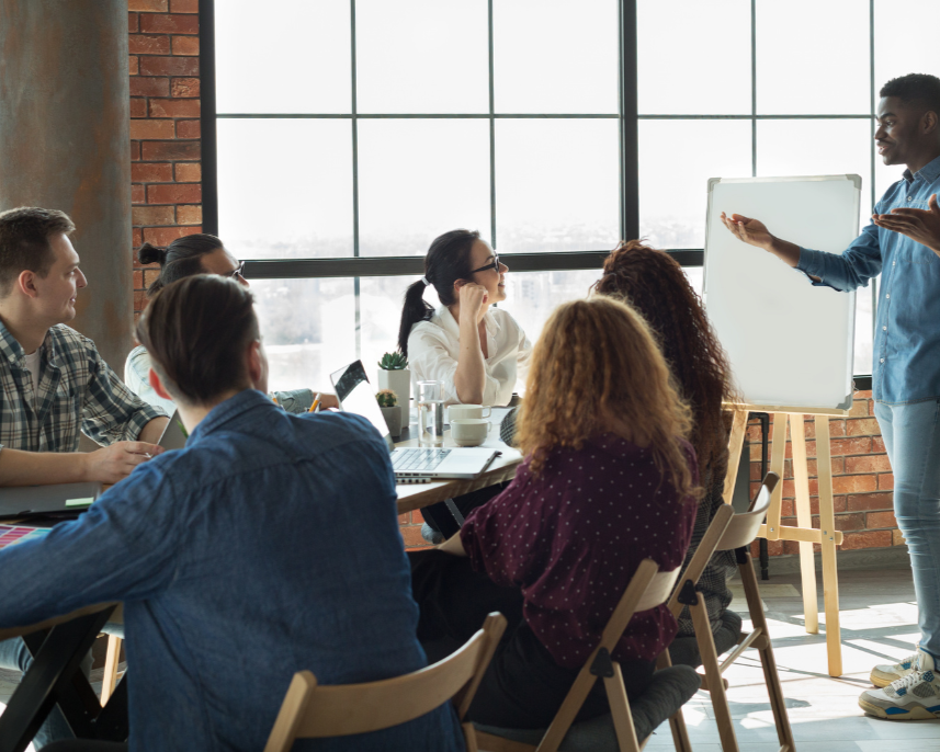 A professional team meeting in a sunlit industrial-style office. A man stands at a whiteboard presenting to a group of seated coworkers.