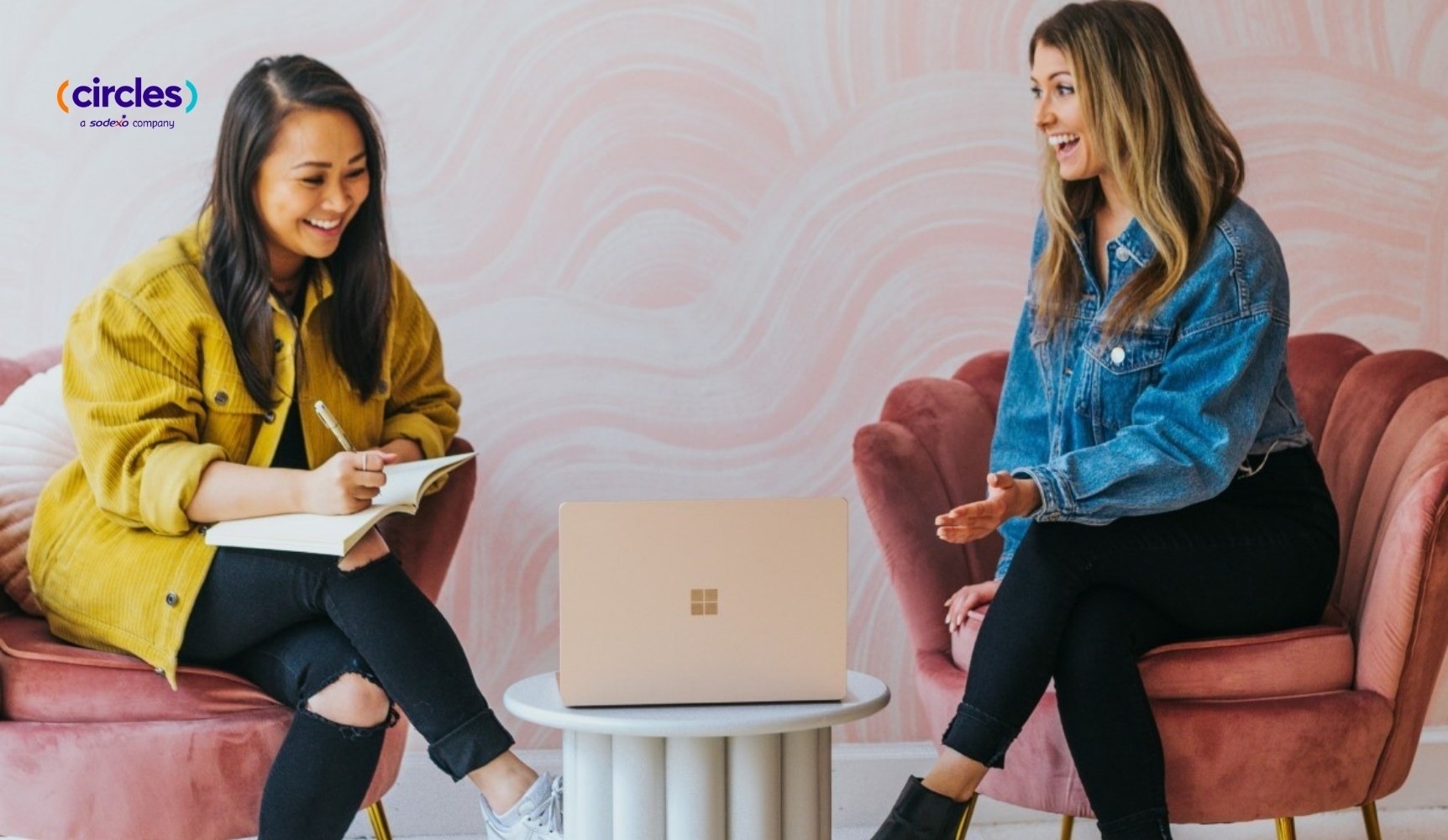 Two women laughing and collaborating during a meeting in a stylish office with a pink marbled wall.
